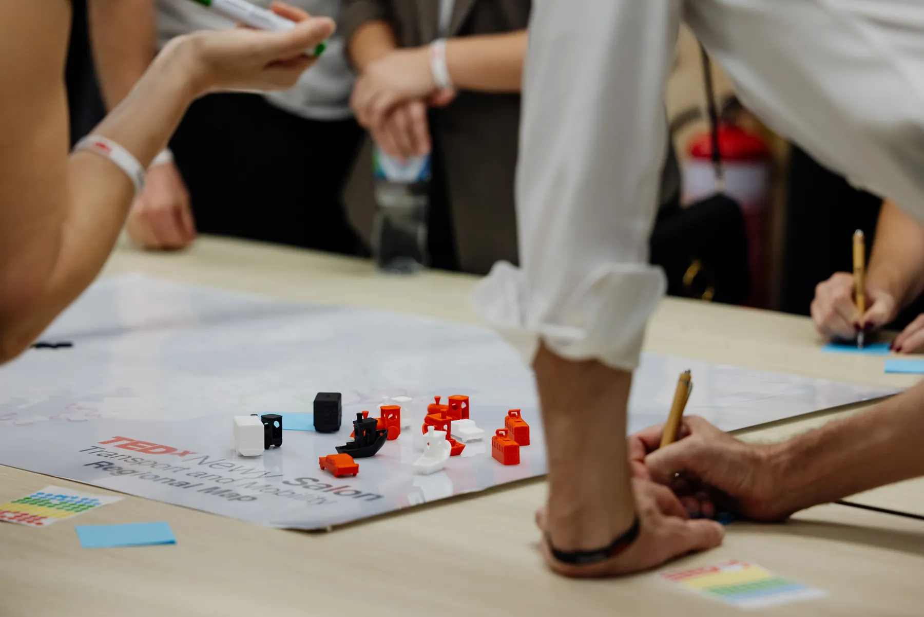 Small coloured blocks and figures arranged on a TEDxNewy Salon table map while people sketch.
