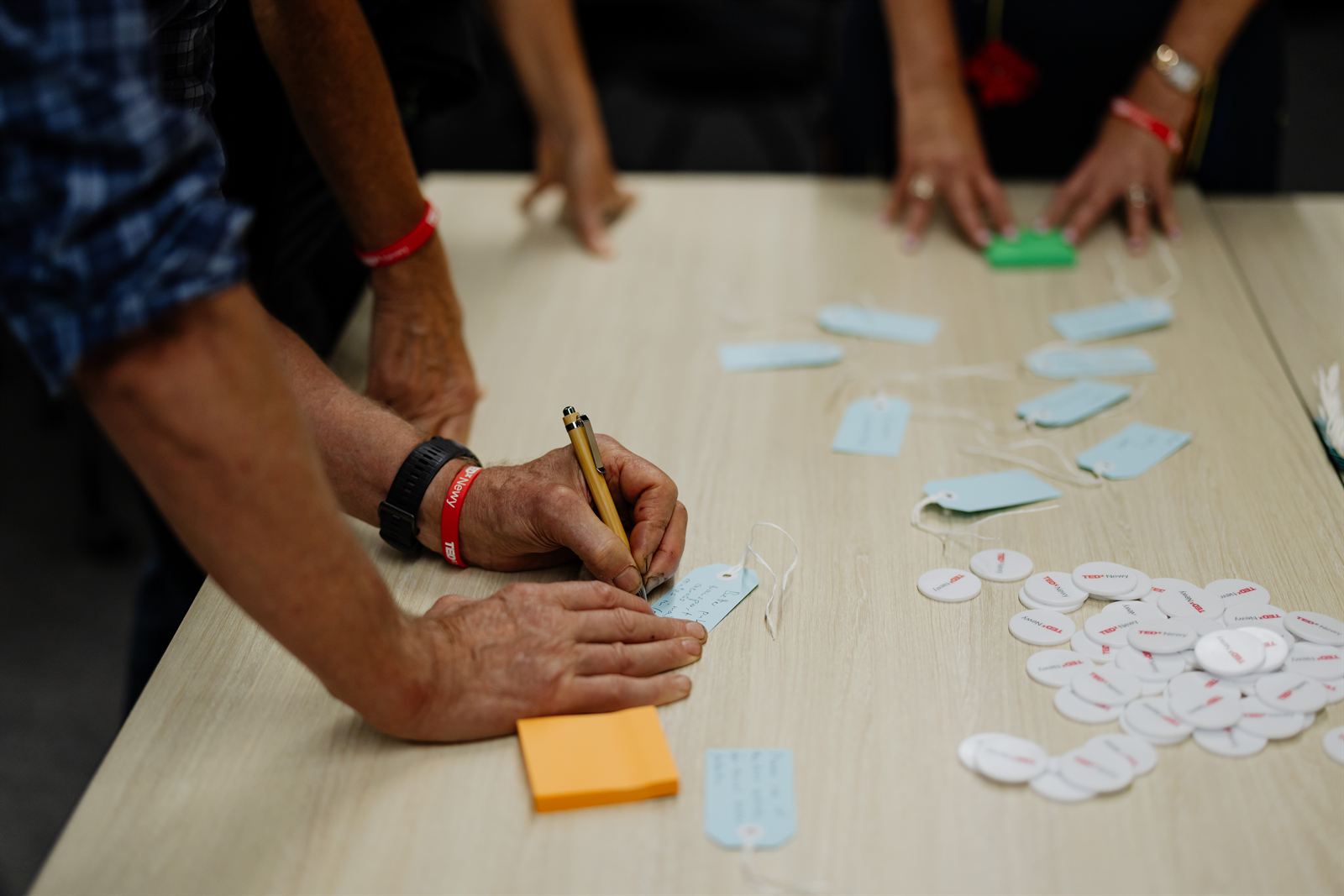 Hands writing on tags during a TEDxNewy workshop, with TEDxNewy badges in the foreground.